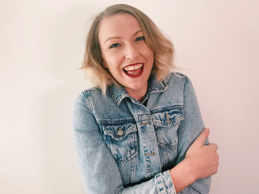 Jen Grieves hugging her arms wearing a denim jacket and smiling laughing to the camera in front of a white background.