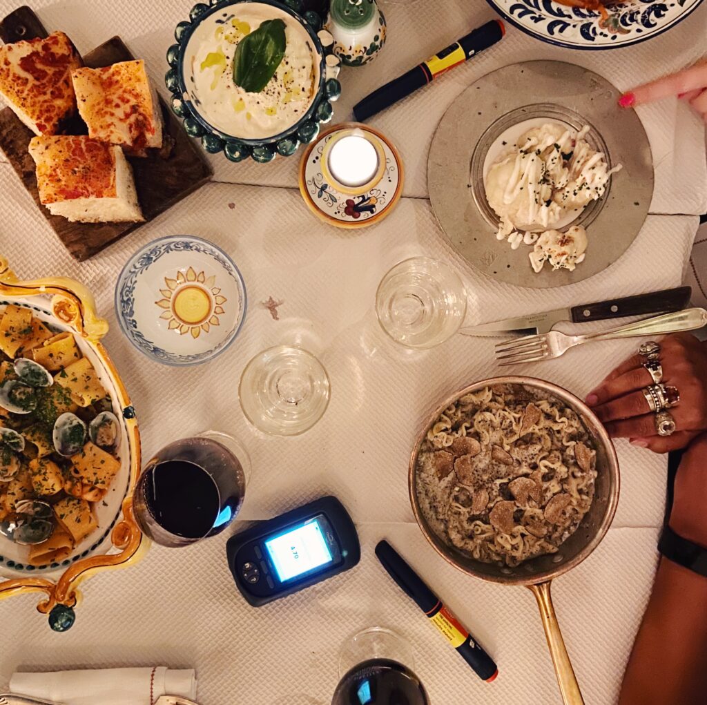 Top shot of a table of food containing pasta, wine, bread and cheese as well as two insulin injections and an Omnipod pump PDM