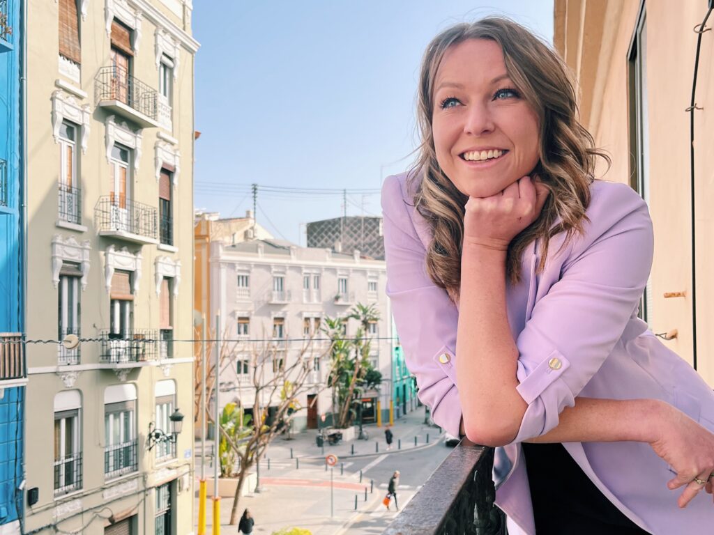 Jen Grieves smiling on a balcony wearing a lilac blazer looking out to the street below