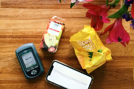 A flat lay of a table showing an Omnipod PDM, a Dexcom graph showing extended overnight hypos, an empty apple juice carton and a packet of dried pineapple