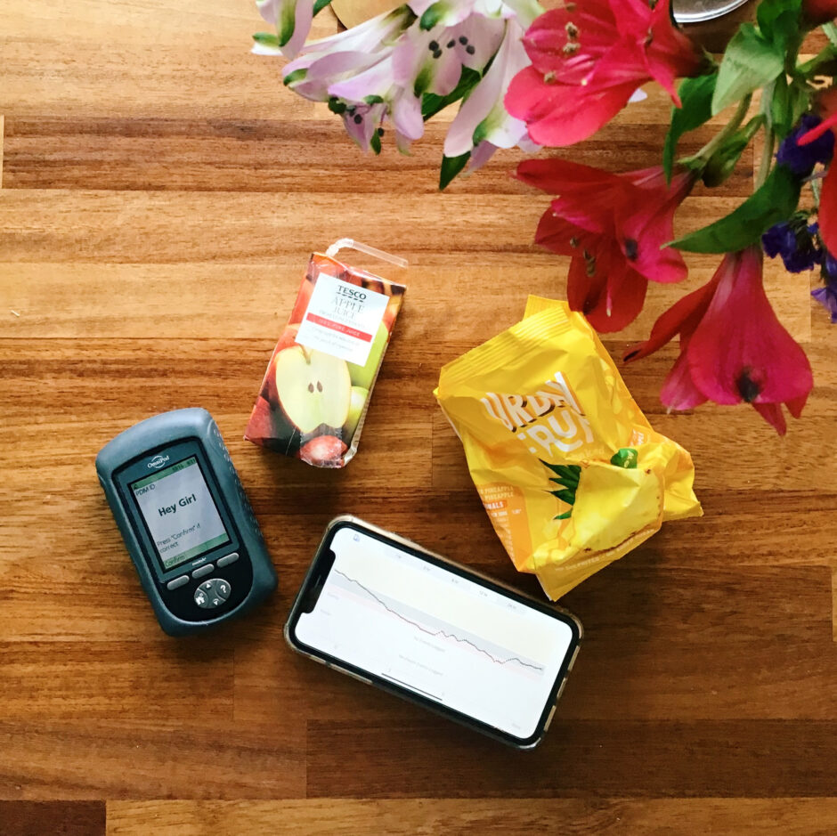 A flat lay of a table showing an Omnipod PDM, a Dexcom graph showing extended overnight hypos, an empty apple juice carton and a packet of dried pineapple