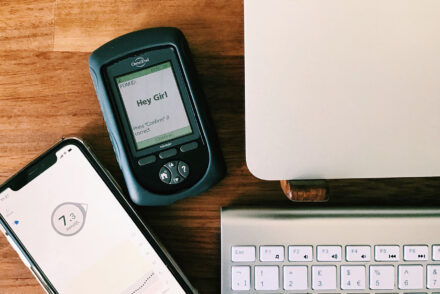 An image of a phone showing a Dexcom CGM reading of 7.3 and an Omnipod insulin pump on a table next to a laptop