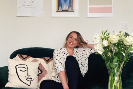A woman (Jen Grieves) sitting on a sofa in front of art with books and flowers on a coffee table